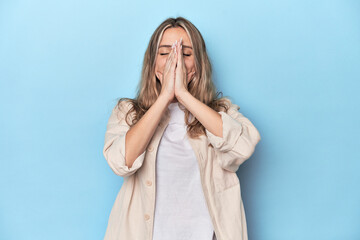 Wall Mural - Blonde young caucasian woman in blue studio holding hands in pray near mouth, feels confident.