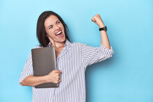 Young Businesswoman Holding Laptop On Blue Raising Fist After A Victory, Winner Concept.