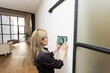 © Angelov - Young woman controlling home with a digital touch screen panel installed on the wall in the living room. Concept of a smart home and mobile application for managing smart devices at home.