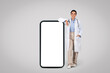 © Prostock-studio - Cheerful latin female doctor leaning on huge smartphone with blank screen, grey background, mockup, full length