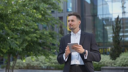 Stylish handsome businessman holding tablet reading email stands on street smiling writes message communicates in social network pensive man browses goods in online store using mobile app. Ukrainian