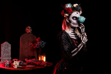 Wall Mural - Glamour woman dressed as catrina skull in studio, wearing traditional santa muerte holy costume with black and white make up or body art. Holding roses and having flowers crown.