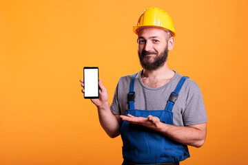 Construction worker holding cell phone with empty screen for advertising in front of camera. Professional builder with hard hat against yellow background in studio shot.