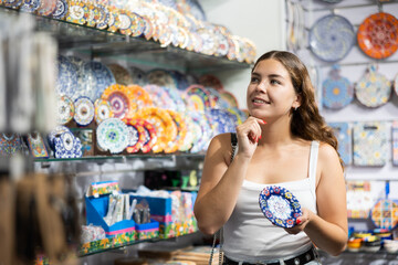 Wall Mural - Tourist girl stopped near window of souvenir shop. Female traveler buyer chooses ceramic decorative wall plate, keepsake