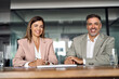 © insta_photos - Smiling Latin mature business man and woman sitting at table in office, portrait. Two happy confident professional mid aged corporate executive leaders company managers wearing suits looking at camera