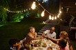 © AnnaStills - Row of lamps illuminating large intercultural family toasting with glasses of wine over table served with variety of food while having dinner