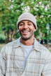 © Jose Calsina - Vertical close up portrait of young hispanic man with a beanie hat smiling and looking at camera outdoors. Front view of latin happy guy standing at park with carefree attitude. Lifestyle concept