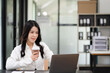 © NINENII - Asian businesswoman sits in a office working on laptop and enjoys a coffee.