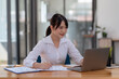 © amnaj - Smiling young happy Asian businesswoman working with laptop and documents in office. Accountant or beautiful asian female worker working happily in the office...