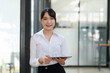 © amnaj - Casual City Lifestyle. Young Asian businesswoman looking at camera holding pen with tablet at office.