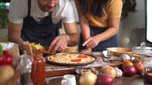 Happy Lovely Couple Enjoy Making A Home Made Italian Pizza Together At Home, Man Adding A Pizza Ingredients And Sauce Over The Pizza.