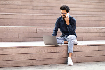 Wall Mural - Indian freelancer young man talking on phone while using laptop