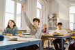 © Maskot - Boy raising hand while sitting with female friend at desk in classroom