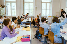 Male And Female Students Raising Hands While Sitting On Chairs In Classroom