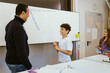 © Maskot - Smiling boy talking to male teacher standing near whiteboard in classroom