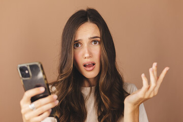 Wall Mural - Mad and shock young brunette beautiful woman using mobile phone isolated on beige background. Yelling unhappy woman hold mobile phone. Disappointed sad upset lady horrified impressed news. WTF. Oh no.