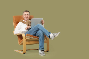 Mature man with laptop messaging in armchair on green background