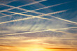 Around 10 airplane vapor trails in the sky in a diagonal pattern against a dark blue sky at sunset with yellow and orange clouds on the horizon.