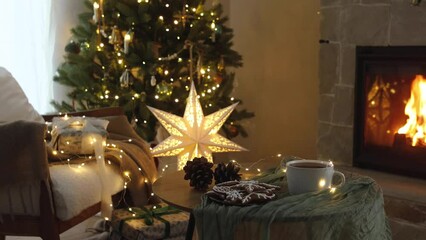 Wall Mural - Cozy christmas eve in festive decorated living room. Stylish gifts, gingerbread cookies and tea cup on table against christmas tree with lights and burning fireplace. Winter hygge footage