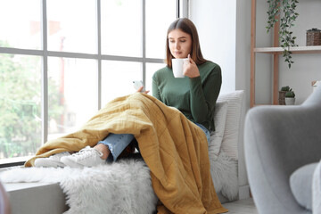 Young woman with cup of hot tea using mobile phone at home