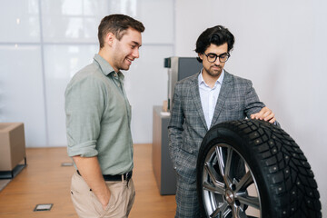 Wall Mural - Front view of salesman in business suit talking about characteristic of tires product to cheerful customer male came to look at assortment represented in car dealership. Concept of auto maintenance.