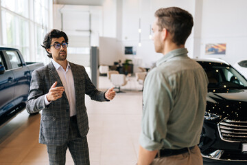 Wall Mural - Portrait of professional male dealer in business suit presenting to handsome young man new model of luxury car. Rear view of unrecognizable male customer choosing new vehicle to buy at showroom.