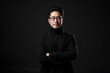 © NaphakStudio - Portrait of a smiling Asian businessman wearing glasses and black suit with looking at the camera in confidently while standing alone in a dark wall background.