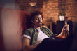 © Marko Geber - Young man relaxing and drinking a cold beer while listening to music on his smartphone on the couch in the living room