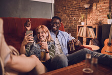 Happy Young Couple Using A Smartphone And Listening To Music While Having Beer On The Couch In The Living Room