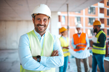 Portrait of satisfied construction site manager wearing safety vest and white helmet at construction site. Young architect watching construction site with confidence looking at camera. 