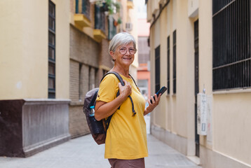 Wall Mural - Happy senior traveler woman in yellow visiting old city of Seville in Spain, using mobile phone looking map directions