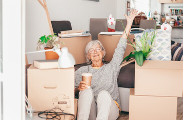 Happy senior woman involved in moving house sitting among cardboard boxes takes a break with a coffee, concept of moving, retirement, new life, buying, renting, apartment, house