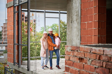 Full length of cheerful man holding daughter and smiling while standing next to wife at construction site. Happy parents with child posing in apartment building under construction.