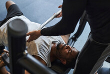 Young Man Doing Bench Press With Help Of Trainer In Health Club