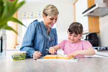 Girl Spreading Butter On Bread By Mother Standing In Kitchen