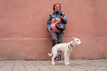 Smiling Woman Standing With Dog And Using Smart Phone In Front Of Wall
