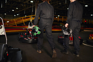 cropped view of two kart racers walking near racing cars and holding helmets, go-kart drivers