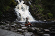 © Aleksandra Iarosh - a young beautiful woman tourist sitting against the backdrop of a waterfall surrounded by forest