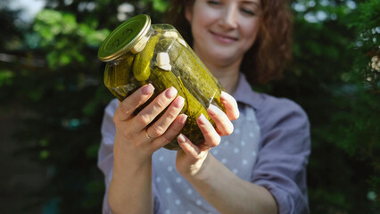 A woman prepares canned vegetables for the winter. On a warm autumn day, the hostess does household chores, picking vegetables for the winter. Pretty woman shows a jar of pickled cucumbers made by