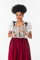 Wall Mural - joyful african american bavarian waitress in dirndl holding empty beer mags and smiling on white