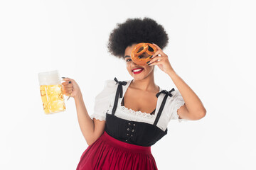 Wall Mural - cheerful african american oktoberfest waitress with mug of beer obscuring face with pretzel on white