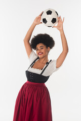 Wall Mural - joyful african american bavarian waitress in dirndl holding soccer ball in raised hands on white