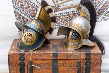 2 ancient roman gladiator helmets on a brown wooden chest with a mosaic image in the background