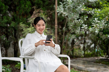 A beautiful and relaxed Asian woman is enjoying her smartphone while relaxing in her home garden.