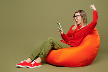 Wall Mural - Full body side view overjoyed young woman she wearing red shirt casual clothes glasses sit in bag chair do winner gesture isolated on plain pastel green background studio portrait. Lifestyle concept.