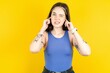Beautiful woman wearing blue tank top covering ears with fingers with annoyed expression for the noise of loud music. Deaf concept.