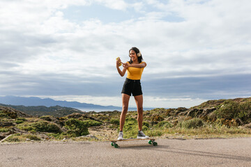 Smiling ethnic woman on longboard and taking selfie