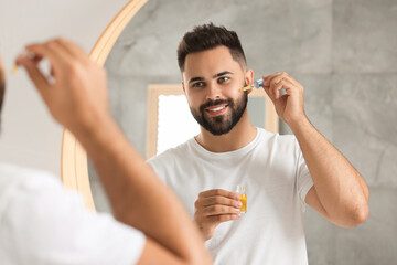 Poster - Handsome man applying cosmetic serum onto his face near mirror indoors