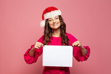Beautiful Christmas Woman In Santa Hat Holding Blank Empty Paper Banner Board On Pink Background, Xmas Portrait