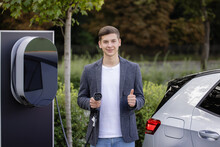 Male Hand Showing Thumbs Up Holding Power Cable Supply Plugged At Electric Car Charging Station. Man Standing Near Electric Charging Station Looking At Camera And Showing Thumb Up